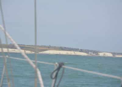Coastline with white chalk cliffs along a calm blue sea, viewed from a sailboat with lines/ropes in the foreground.