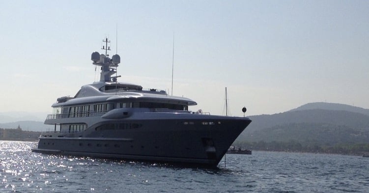 Large luxury motor yacht anchored near a coastline on calm water with hills in the distance.