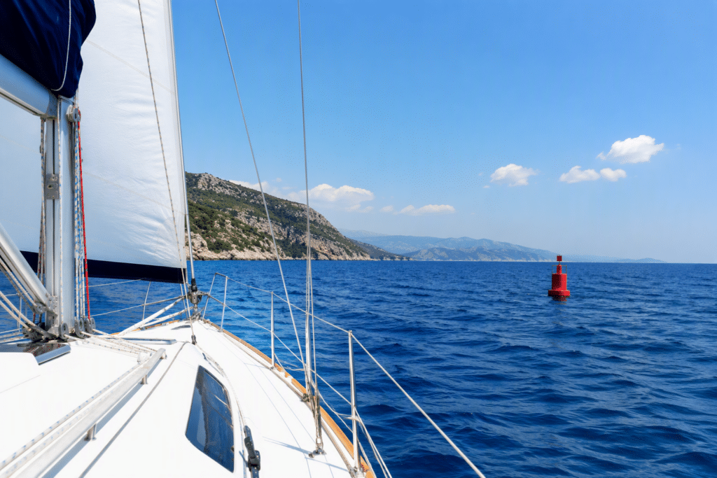 Sailboat deck with rigging and mast, blue sea, rocky coastline in the distance, and a red buoy on the water under a clear sky.