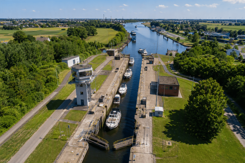 Aerial view of a canal lock with boats in the lock chamber, a tall control tower, and green fields on both sides.